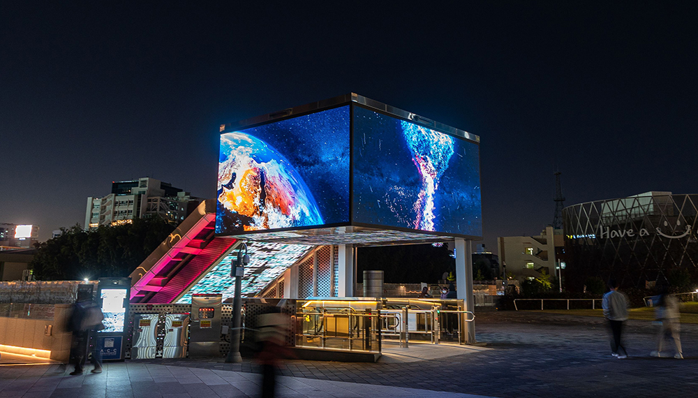 Night scene of a large illuminated cube screen displaying Earth, mounted above a pedestrian plaza with pink-lit stairs and passersby nearby.