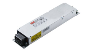 Metal IEC power supply module with mounting flanges and label stickers, viewed from end with output connector visible at front.