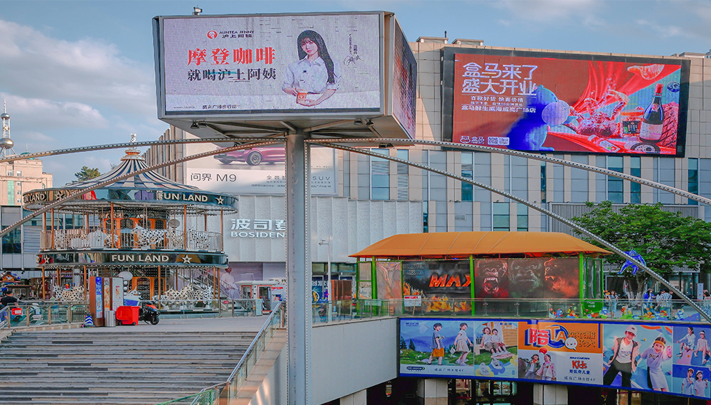 Led-Display-Panels - LED Controller Card Store for LED Display Module, LED Display Parts A bright outdoor shopping complex with large digital billboards, a two-level structure, and a colorful fun‑land ride area on the left side.