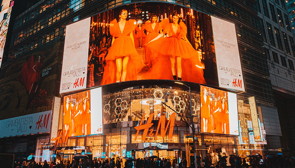 Led-Display-Screen-for-Advertisement - LED Controller Card Store for LED Display Module, LED Display Parts Nighttime urban shopping district with large illuminated billboards showing models in orange dresses above an H&M storefront with crowds below.